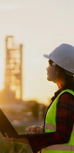 A woman in a hard hat and safety vest standing in a field holding a laptop at sunset.