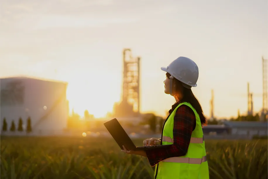 A woman in a hard hat and safety vest standing in a field holding a laptop at sunset.