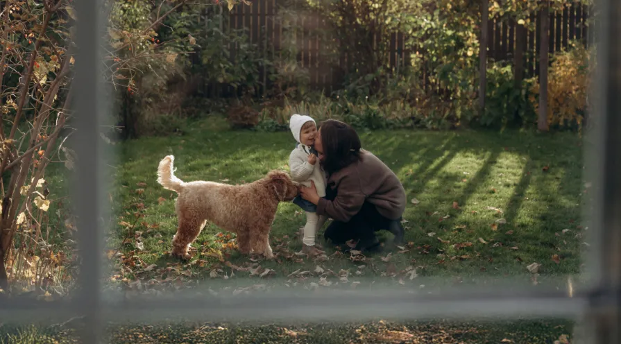 A woman crouching in a sunlit garden, holding a toddler in a white outfit, while a fluffy dog stands nearby.