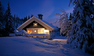 Cozy cabin warmly lit in a snowy forest at dusk surrounded by snow-covered trees.