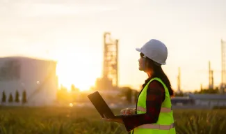 A woman in a hard hat and safety vest standing in a field holding a laptop at sunset.