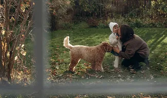 A woman crouching in a sunlit garden, holding a toddler in a white outfit, while a fluffy dog stands nearby.